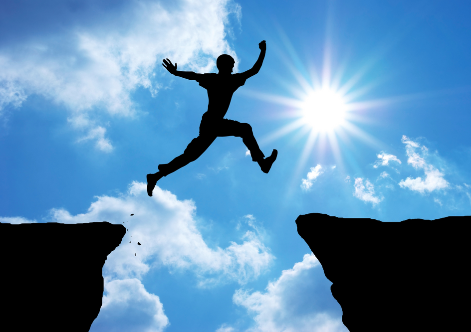 Silhouette of a person jumping over a gap between two cliffs against a bright blue sky with clouds and the sun.