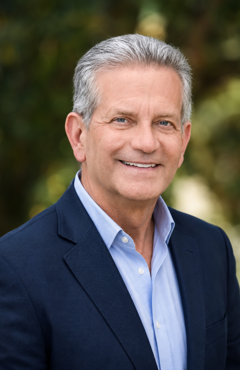 A smiling middle-aged man with gray hair and blue eyes, wearing a navy blazer and light blue shirt, standing outdoors with blurred greenery in the background.