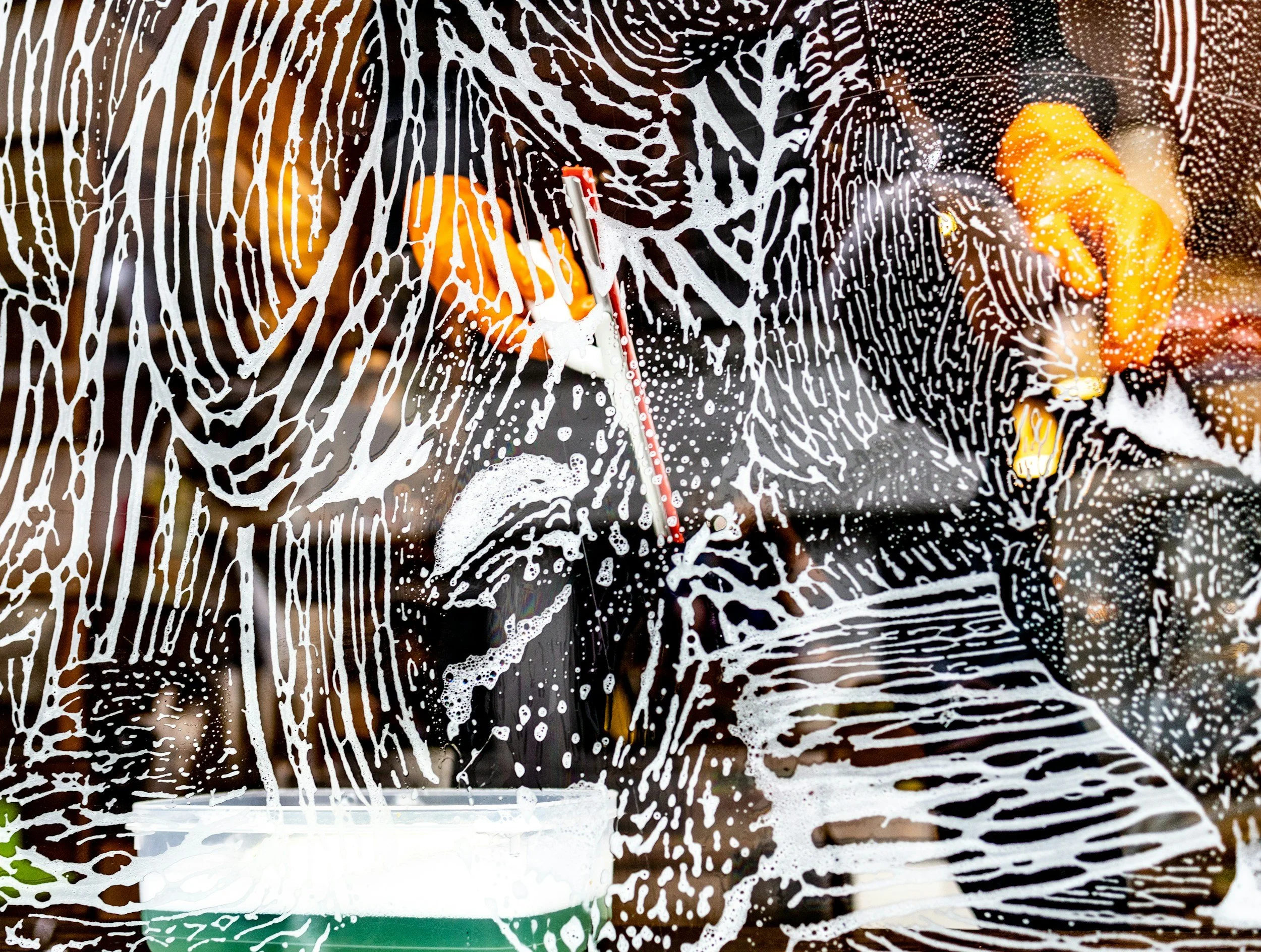 A person cleaning a glass window with a squeegee, covered with soap and foam, creating streaks and patterns.