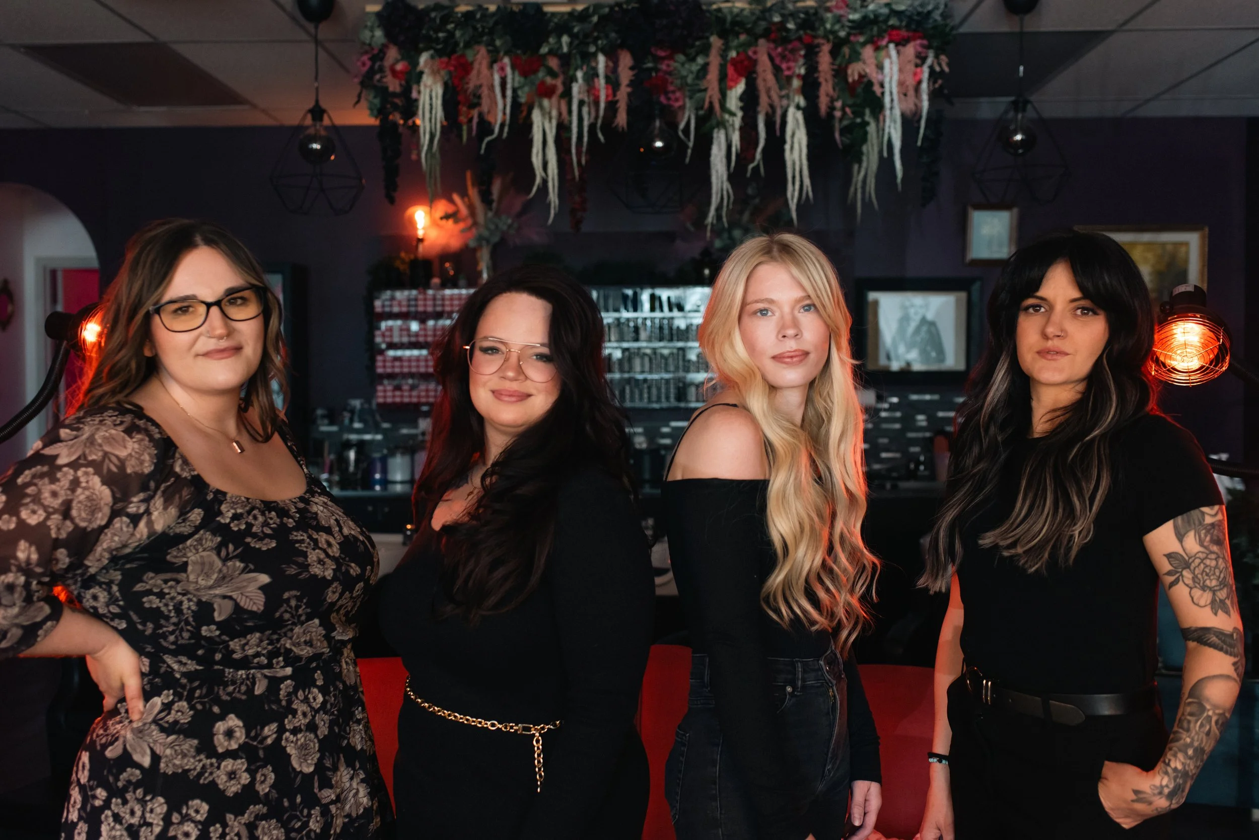 Four women standing together indoors, with dark-colored walls and hanging decor, including flowers and geometric light fixtures, with red lighting and a bookshelf in the background.