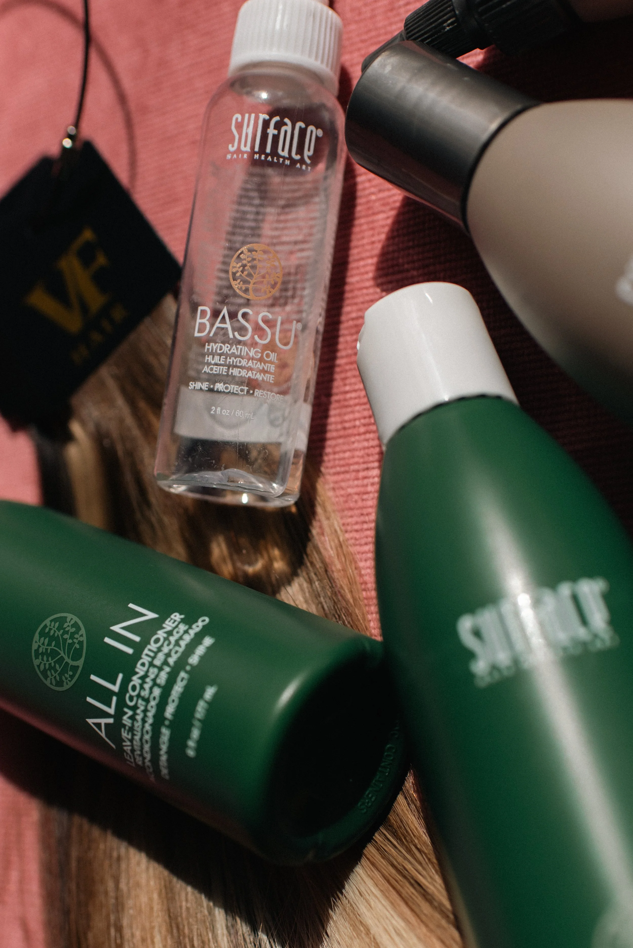 A variety of skincare and haircare products on a reddish-brown textured surface. Visible items include a transparent bottle labeled 'SURFACE Bassu Hydrating Oil,' a green tube with white text, a white spray bottle, and a black container with a black lid.
