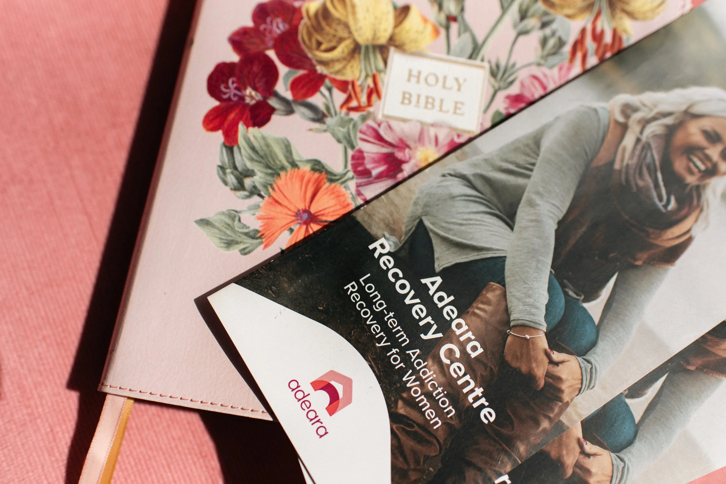 Close-up of a pamphlet featuring flowers, a Bible, and a smiling woman. The pamphlet promotes an addiction recovery program led by Rev. Andrea Recco. The background includes pink and black sections.