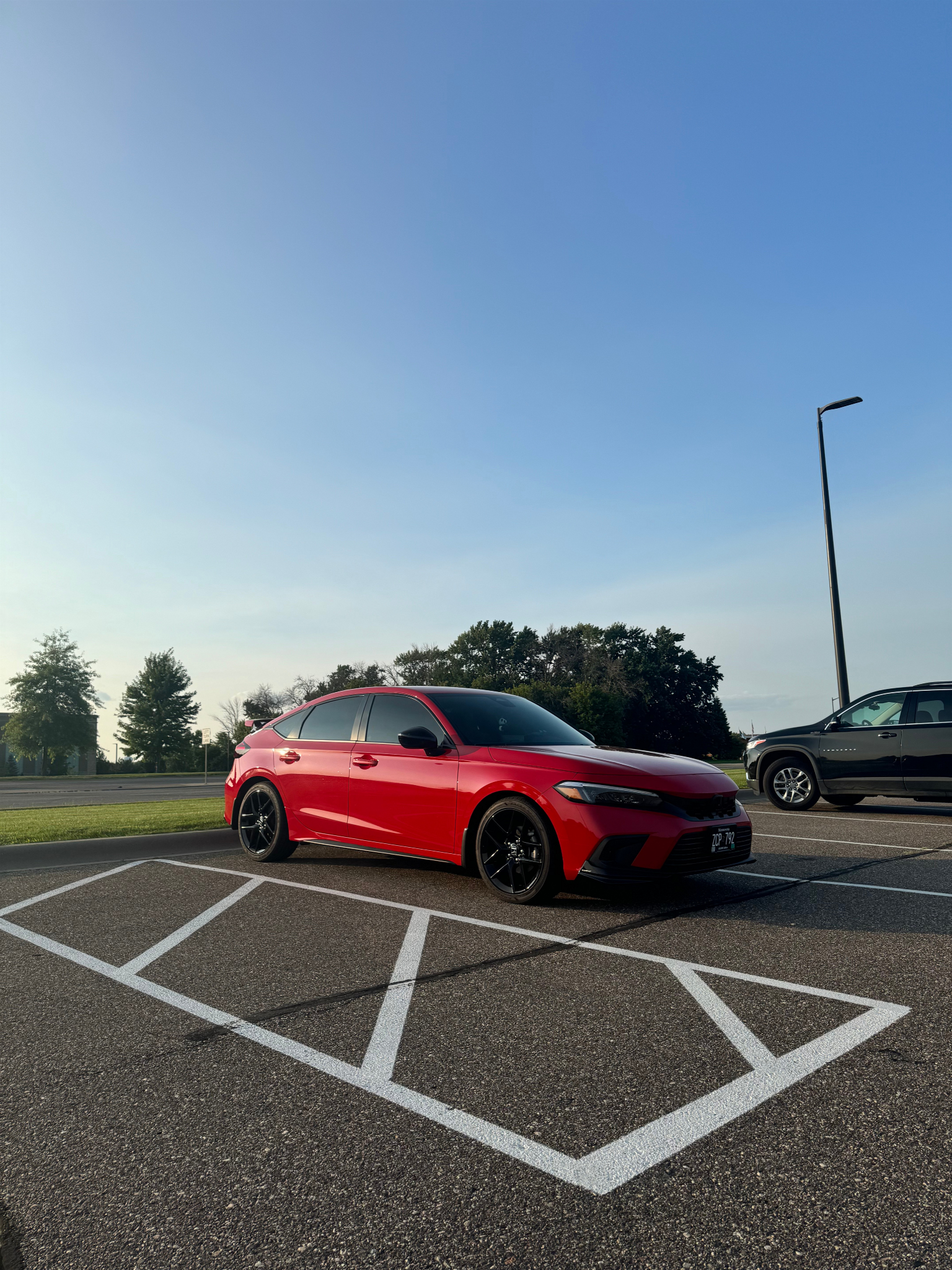 A red car parked in a marked parking space next to a black SUV, with trees and a clear blue sky in the background and a tall streetlight on the right side of the image.