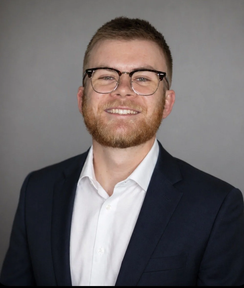 Portrait of a young man with light brown hair, beard, glasses, wearing a suit and white shirt, smiling against a plain gray background.