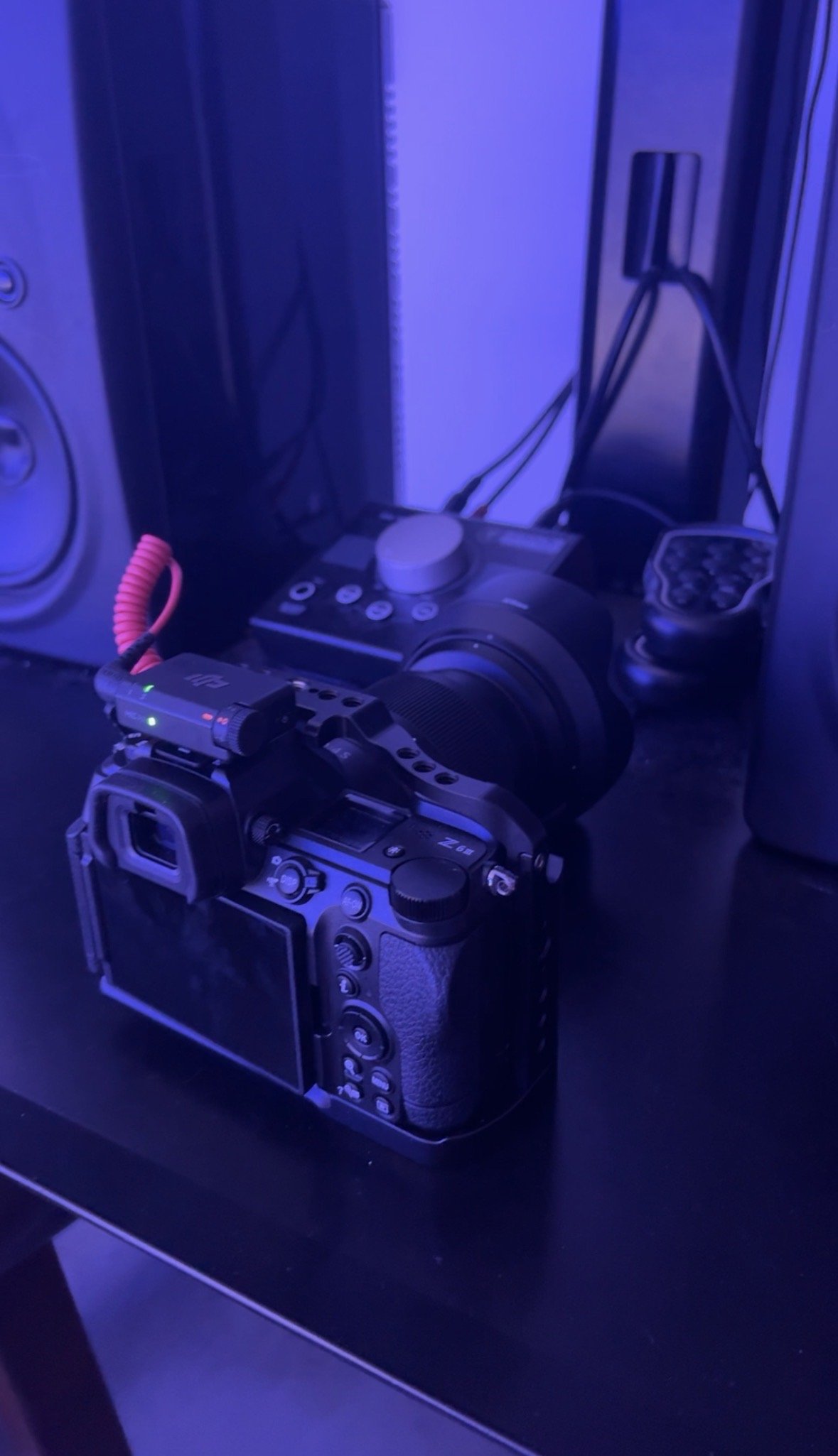 A camera with a grip is placed on a dark surface, with a printer, a controller, and cables in the background under blue lighting.