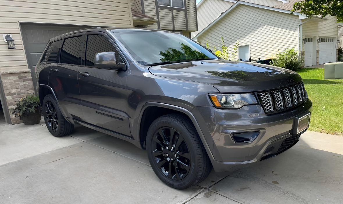 Gray Jeep Grand Cherokee parked in driveway near house with garage and greenery.