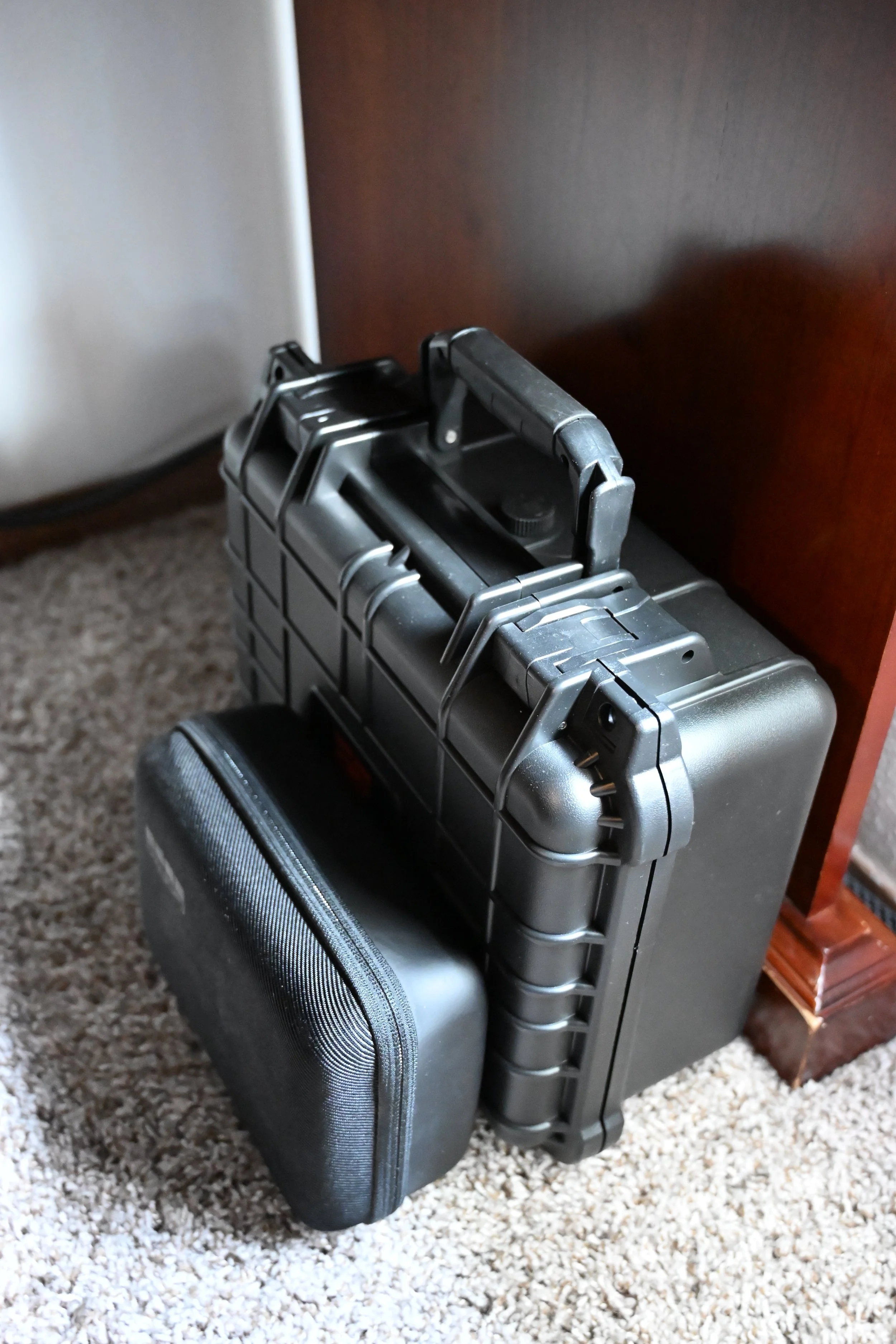 A metal briefcase with a black textured surface, placed on a carpeted floor next to a small black hard case, with a luggage handle and a partial view of a silver appliance and a wooden furniture piece in the background.