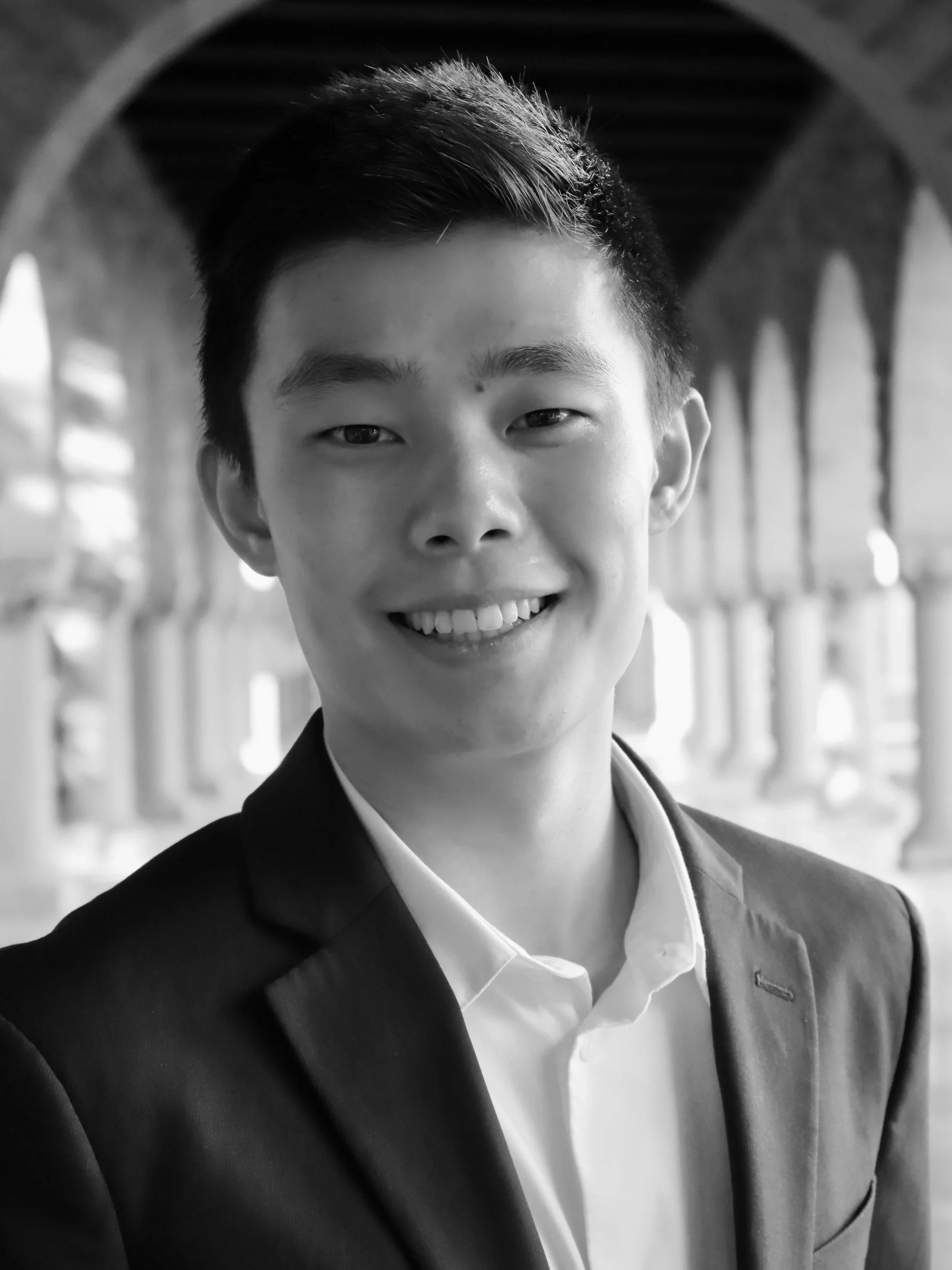 A young man in a suit and white shirt smiling at the camera, with the arched structure of Stanford University in the background.