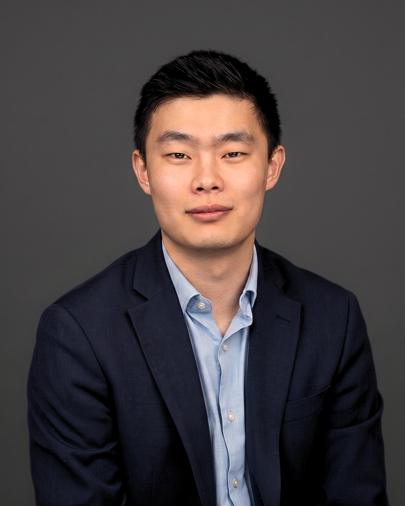 A professional headshot of a young Asian man with black hair, wearing a dark suit jacket and a light blue dress shirt, against a gray background.