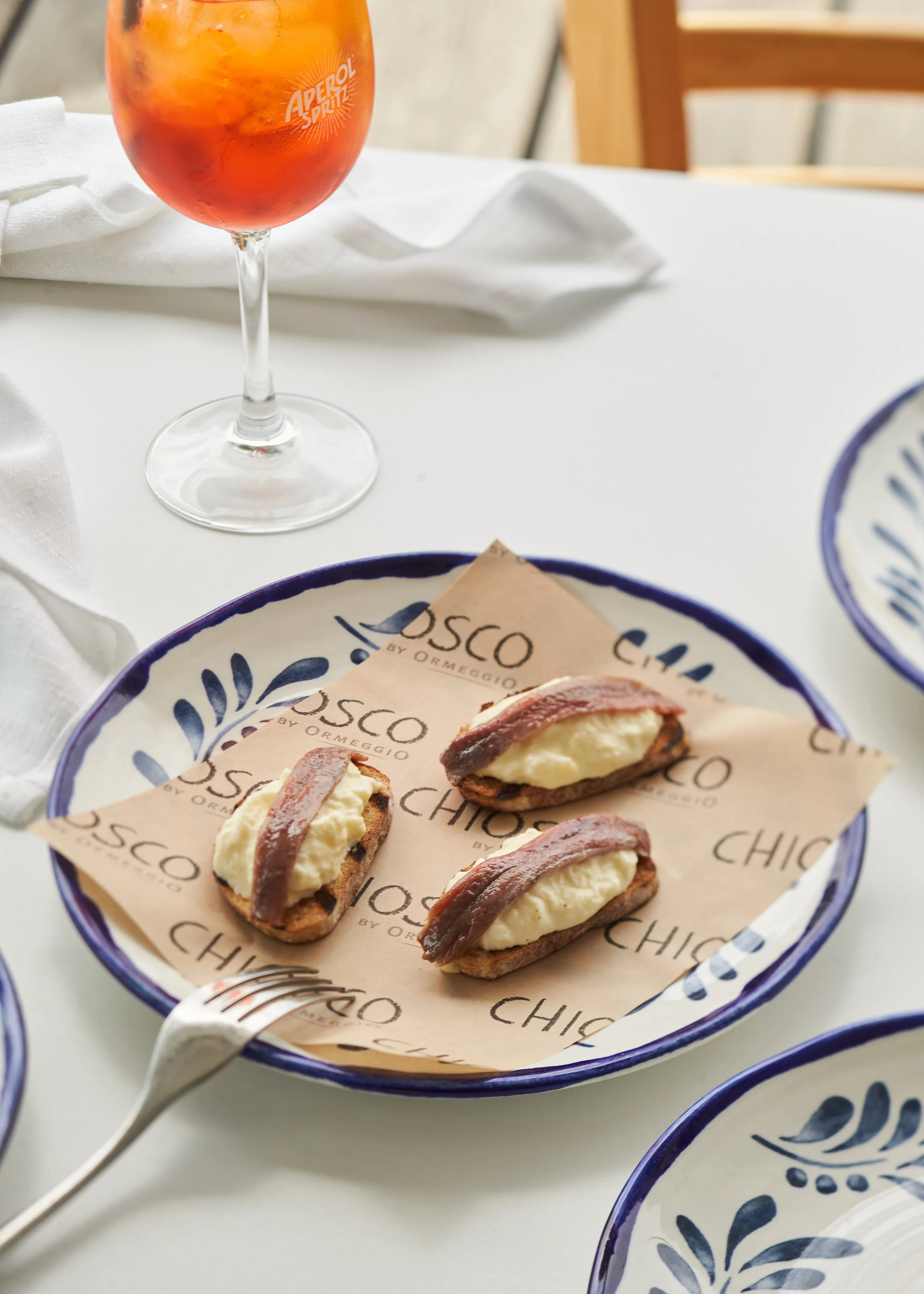 Three crostini toppings with creamy cheese and bacon slices on a blue and white patterned plate, placed on a white tablecloth with a fork nearby. In the background, there is a glass of red-orange beverage with ice and a white napkin.