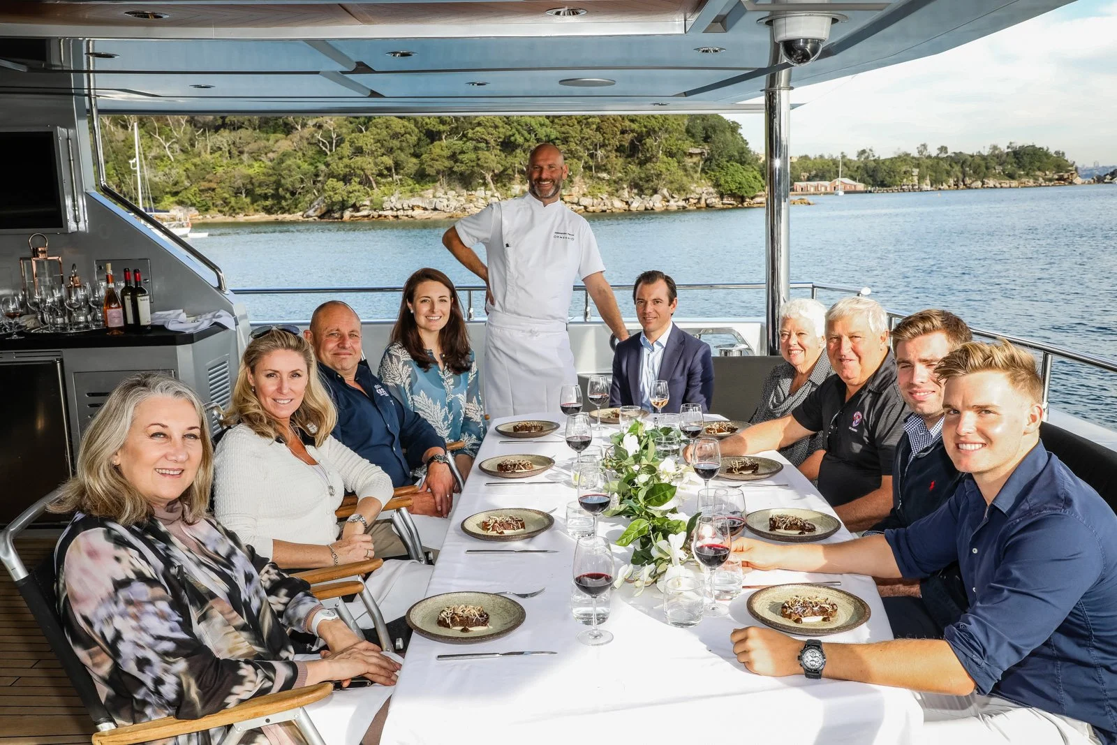 Group of people dining on a boat with a chef standing behind them, scenic water and trees in the background.