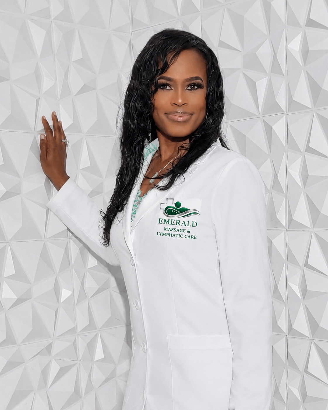 A woman with long black curly hair in a white coat standing against a white textured wall, smiling, with a logo that reads 'Emerald Massage & Lymphatic Care' on her coat.