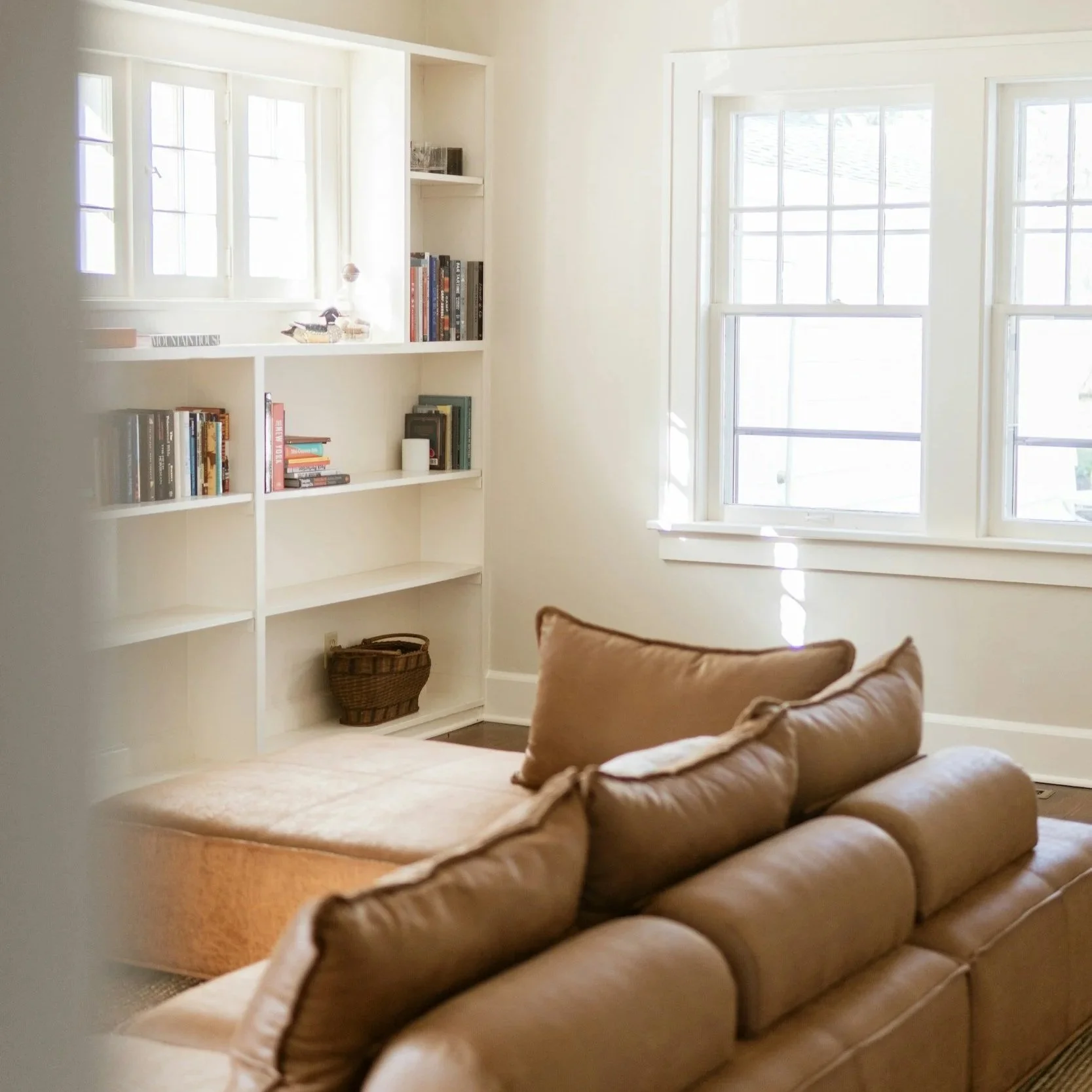 Bright living room with large windows, white built-in shelves filled with books, and a tan leather sofa with pillows.