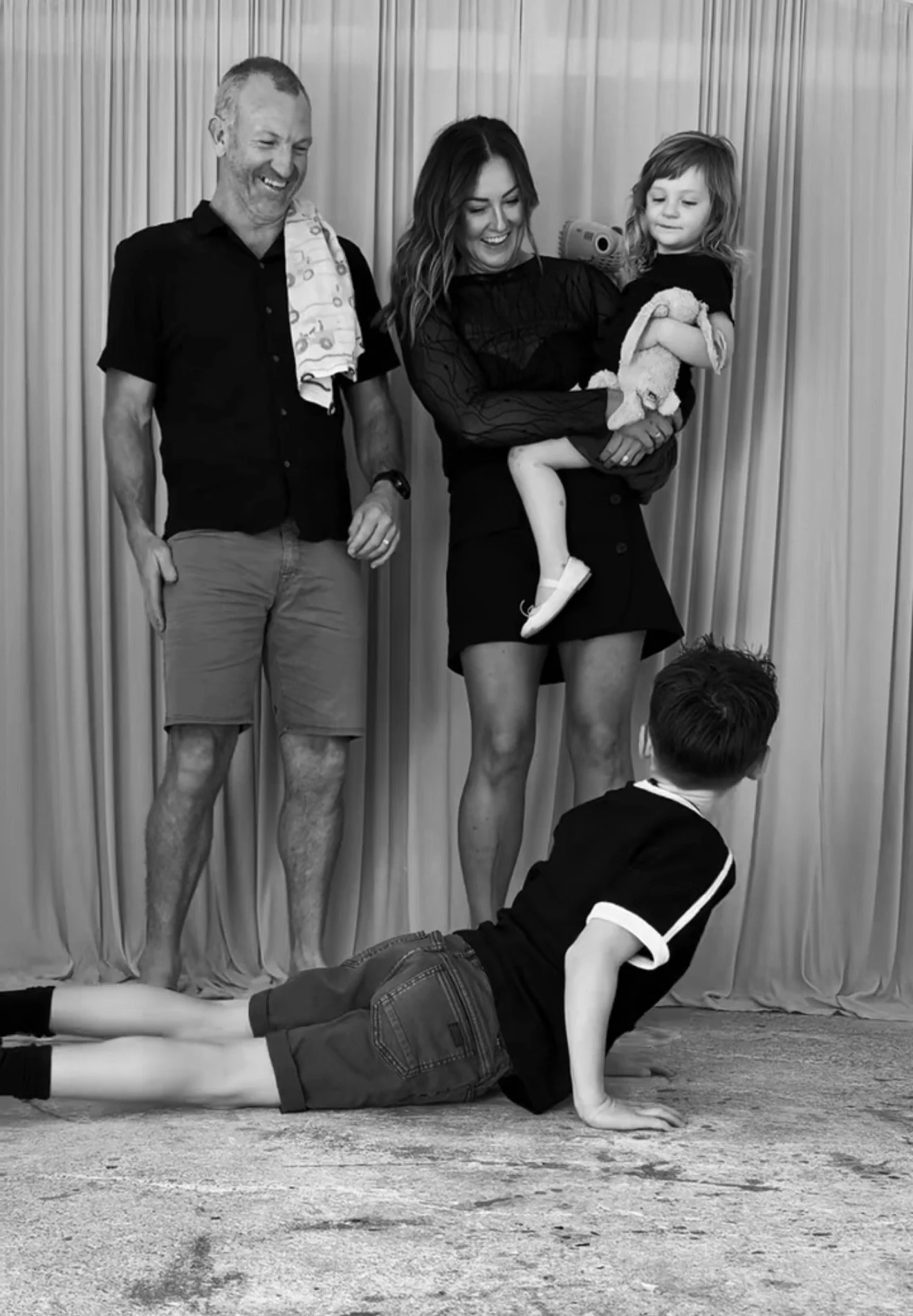 A woman and a man standing behind a boy doing push-ups on the floor, with a girl holding a stuffed animal in front of them, all smiling, in front of a curtain.