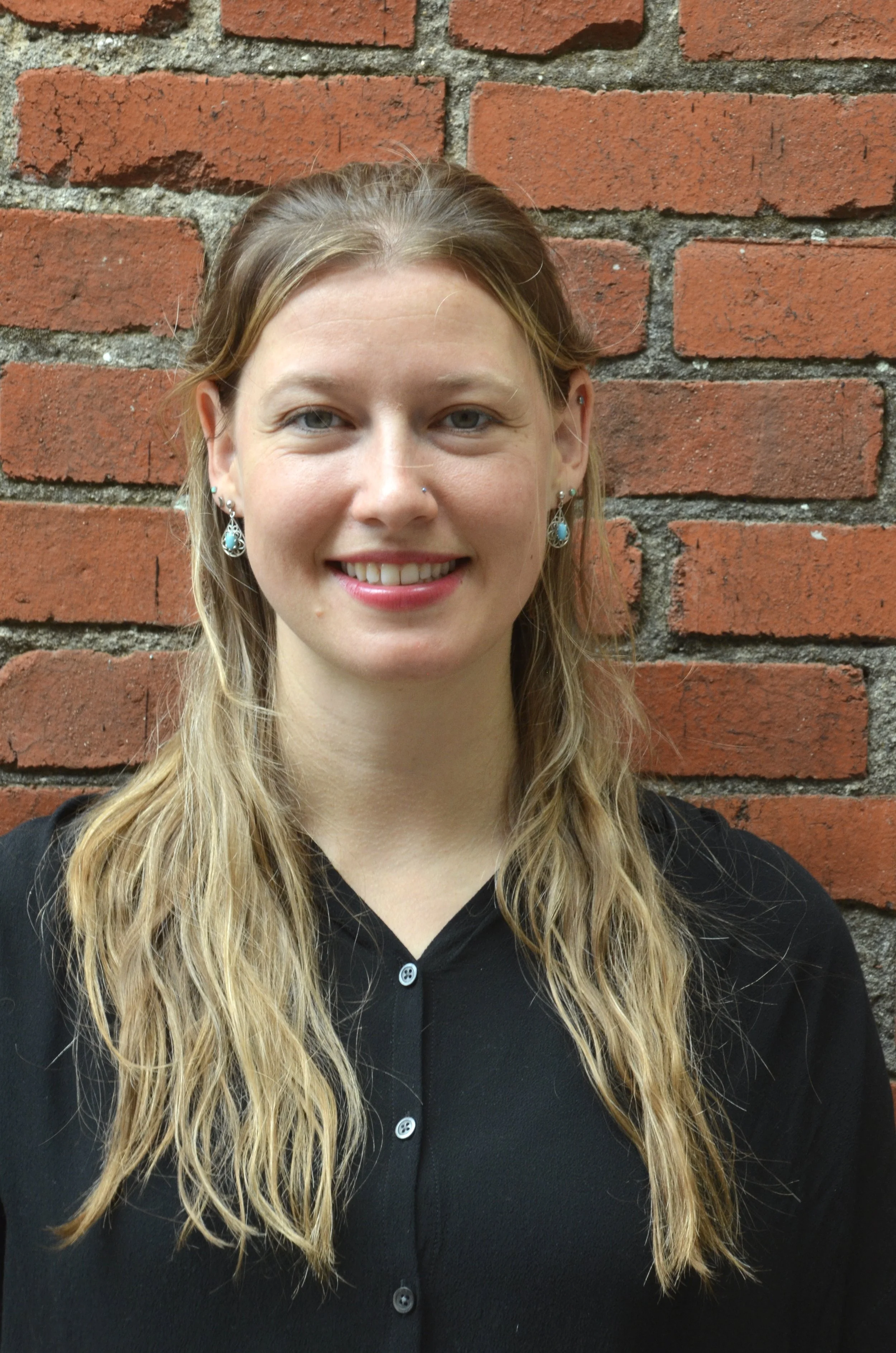 A woman with long wavy blonde hair, wearing earrings with turquoise stones, smiling, standing in front of a red brick wall.
