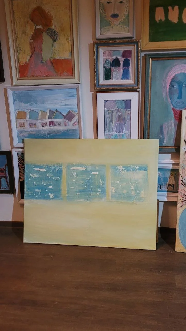 A collection of various colorful paintings, with one large abstract painting of a seascape in the foreground, displaying a yellow sky, blue water, and structures along the shoreline. The background features multiple artwork hung on a wall, including portraits, landscapes, and abstract pieces in different frames.