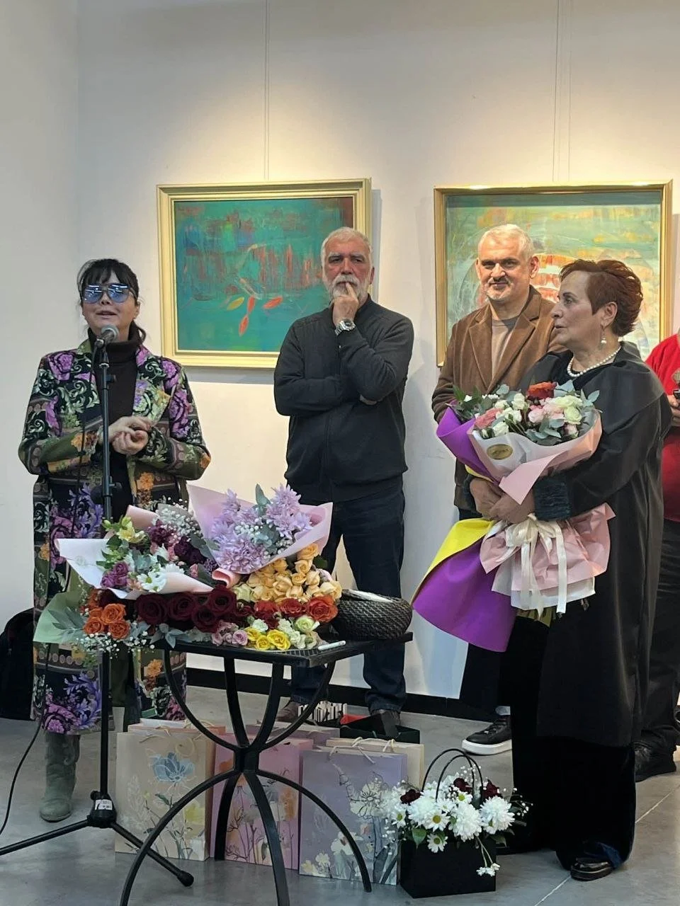People stand in an art gallery during a celebration, holding bouquets of flowers, with paintings on the wall behind them.