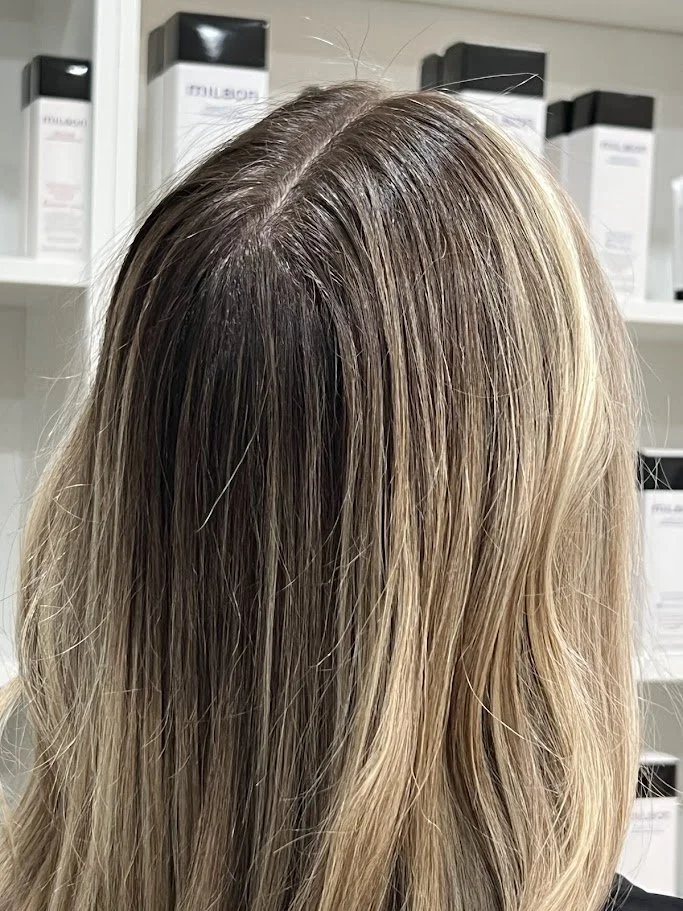 Back of a person's head with blonde hair showing natural roots and straight strands, in a hair salon with shelves and hair products in the background.