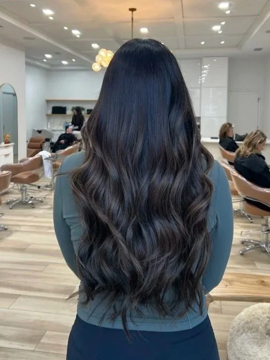 Back of woman with long, wavy, dark hair in a salon with salon chairs and clients in the background.