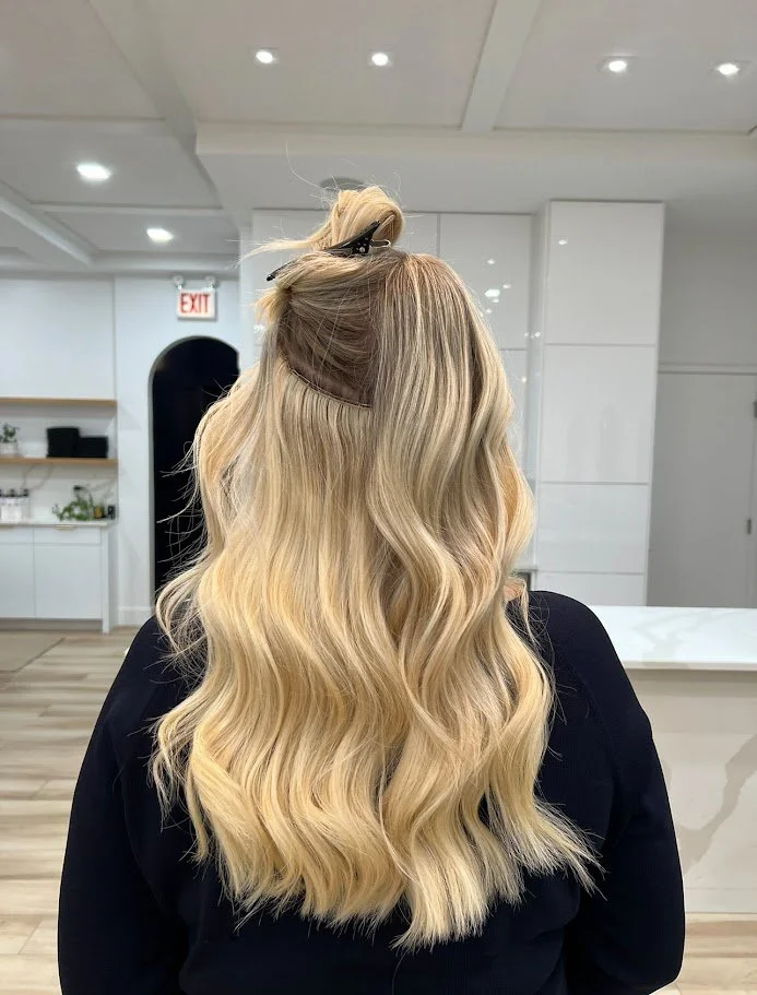 Back view of a woman with long, wavy blonde hair in a salon.