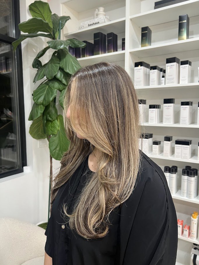 Woman with long, wavy, light brown hair with subtle highlights in a salon or spa setting, standing next to a large green leafy plant and shelves filled with hair or skincare products.