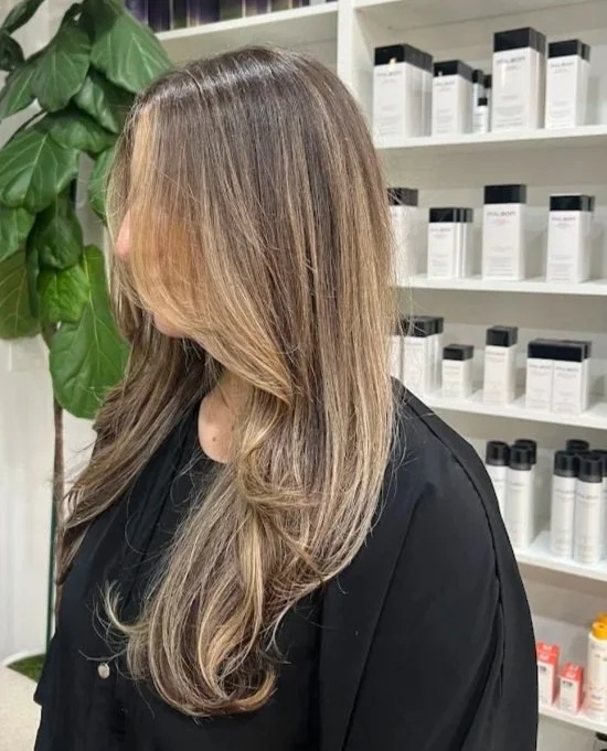 Side view of a woman with long, layered, light brown hair with blonde highlights, standing in front of white shelves filled with hair and beauty products, in a salon or store.