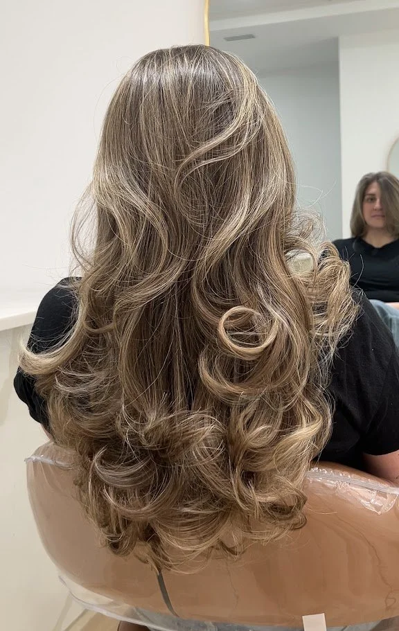 Back view of a woman with long, voluminous, curly blonde hair sitting in a salon chair.