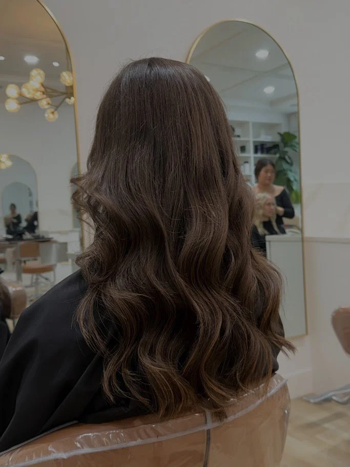 Back view of a woman with long, wavy brown hair sitting in a salon chair, with mirrors and other clients in the background.