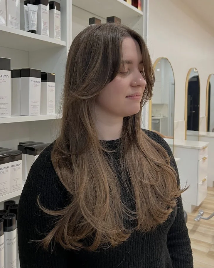 A woman with long, wavy brown hair and a nose piercing, standing in a salon with shelves of hair products in the background.