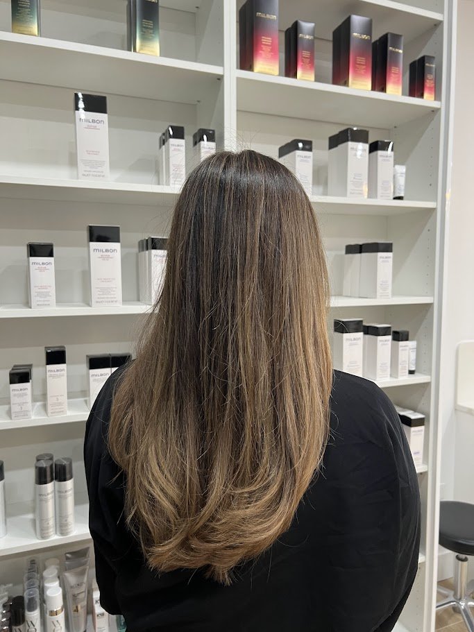A woman with long, straight, light brown hair standing in front of shelves stocked with hair care products.