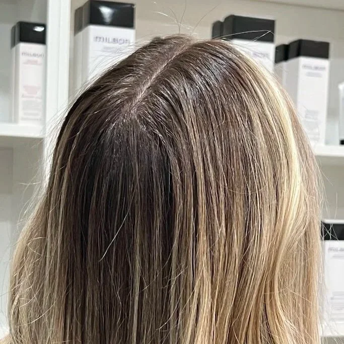 Close-up of a person's blonde hair showing roots and highlights in a hair salon with product bottles on shelves in the background.