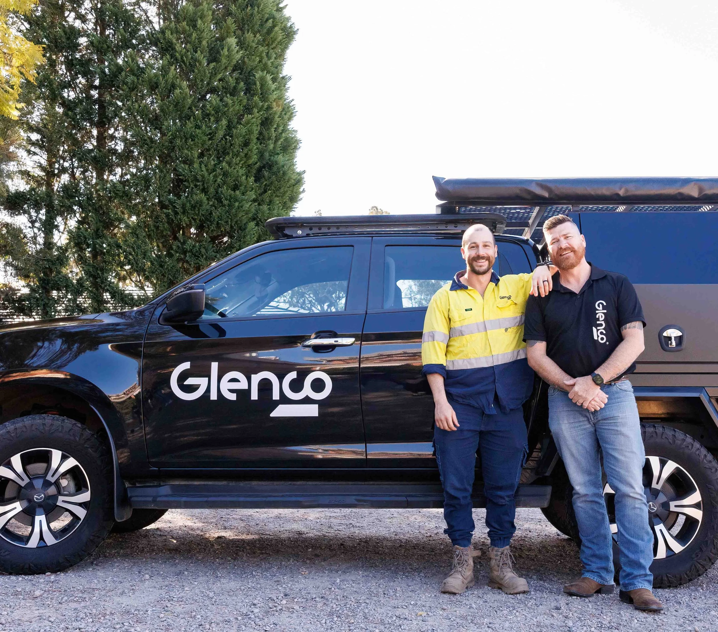 Two smiling men standing next to a black Glenco vehicle, with a large tree in the background, on a gravel surface during daytime.