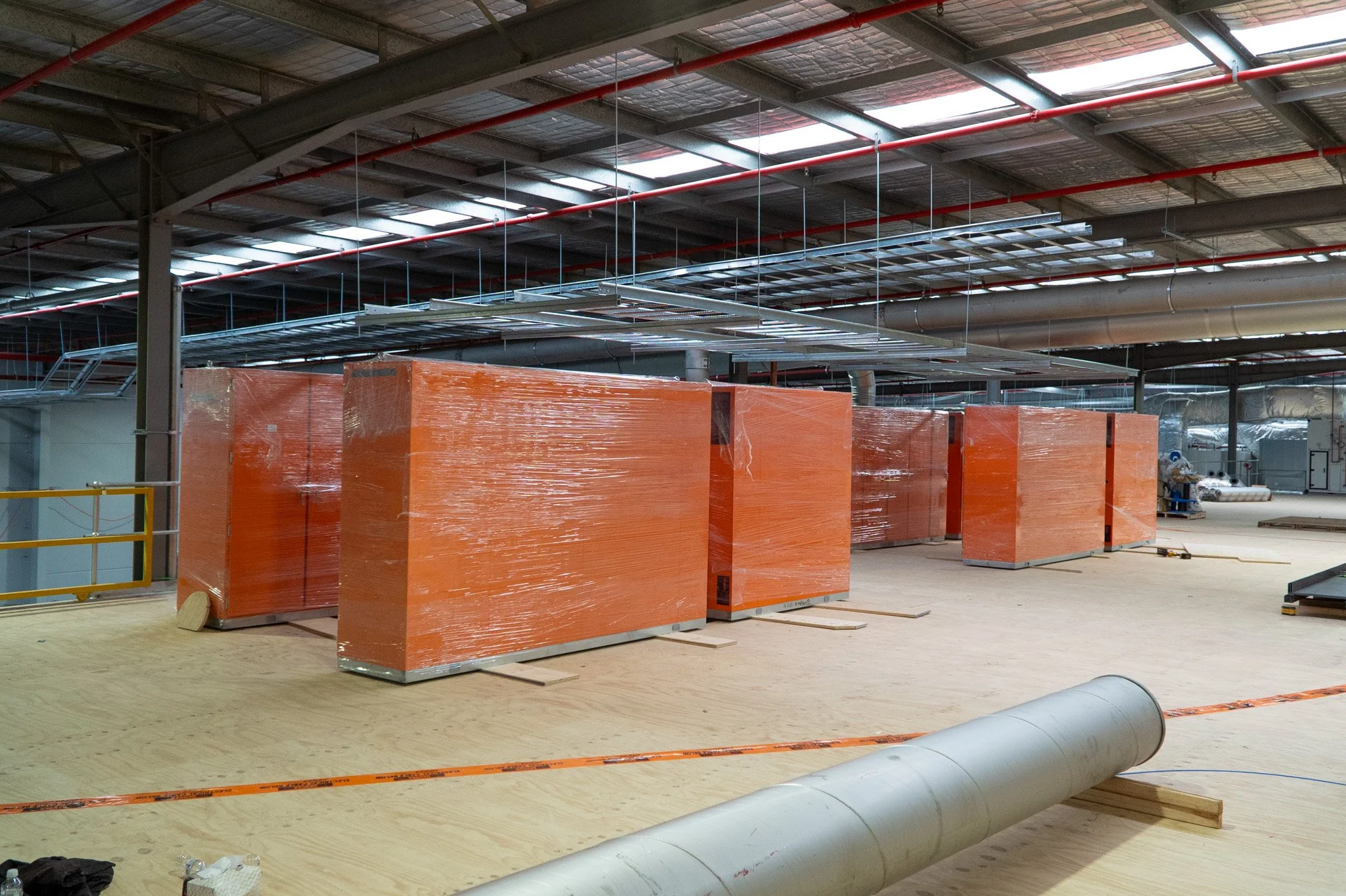 Construction site interior with large orange equipment, metal pipes, and ductwork, and a wooden floor in progress.