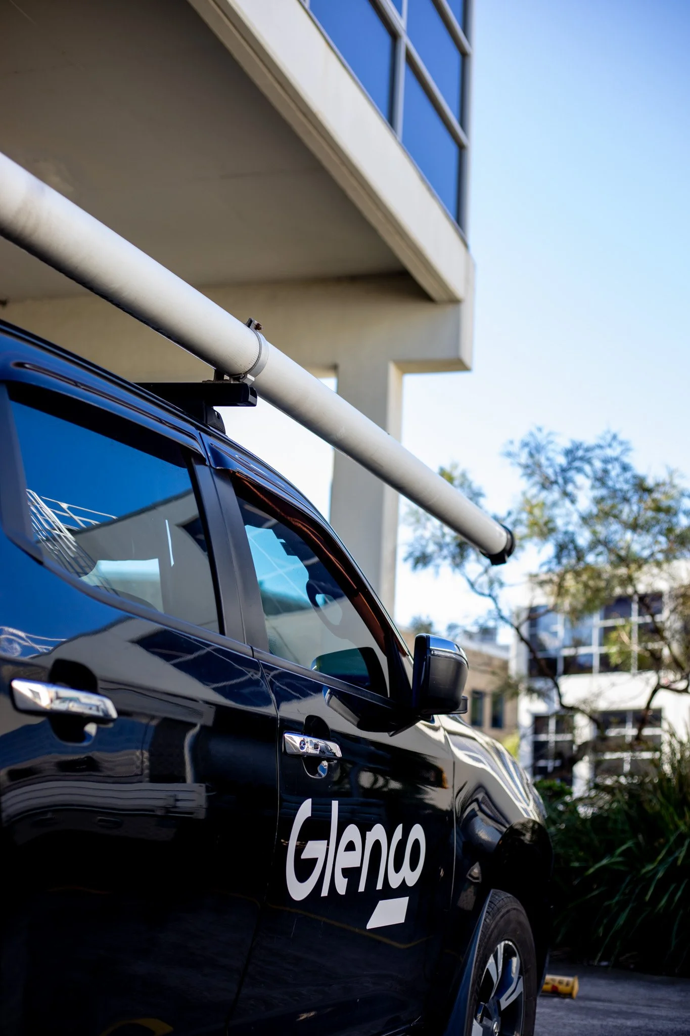 Black vehicle with 'Glenco' written on the door parked outside a modern building with blue-tinted windows, piping on the vehicle roof, and trees in the background under a clear sky.