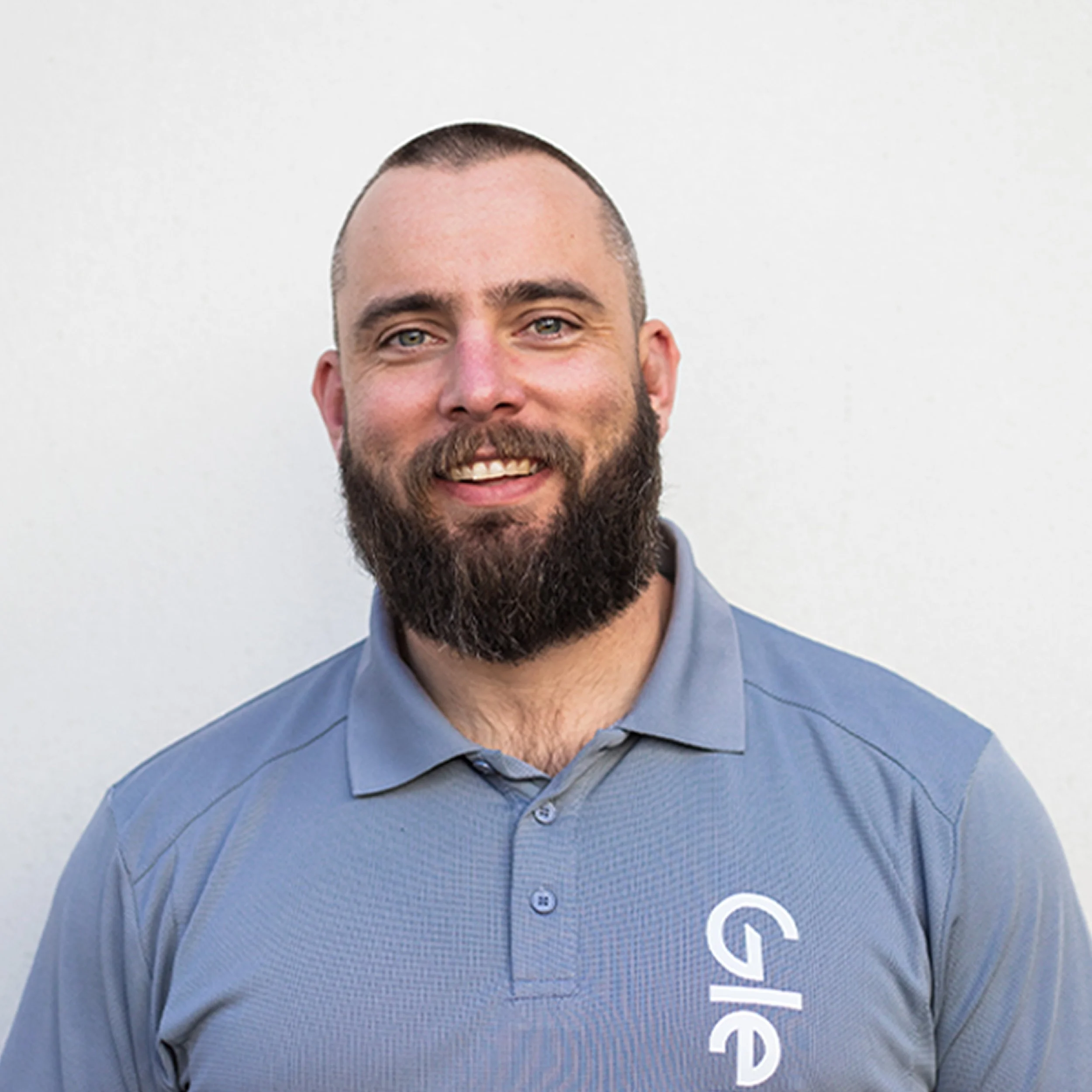 Close-up of a smiling man with a beard in a light blue collared shirt with white text.
