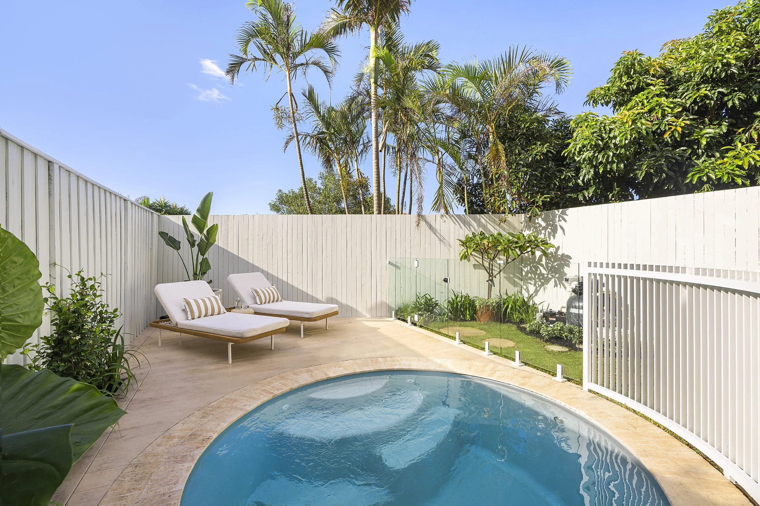 A backyard swimming pool with two lounge chairs on a wooden deck surrounded by lush green plants and trees, with a white fence and clear blue sky in the background.
