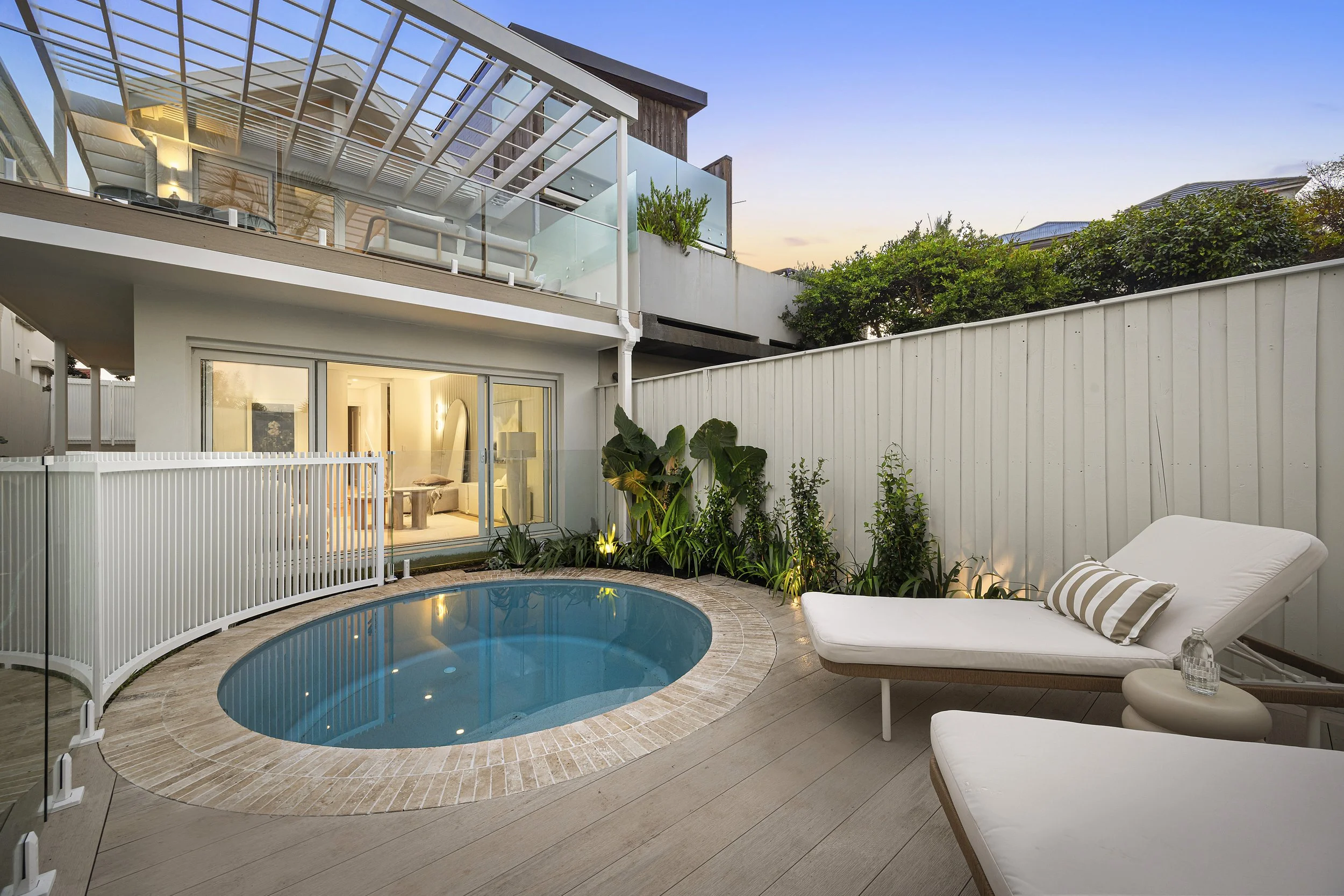 A backyard patio with a small, circular swimming pool surrounded by a light-colored brick edging and outdoor lounge chairs with striped cushions. The area is enclosed by white fencing and garden plants, and there is a modern, multi-story house with large windows and a balcony overlooking the yard at sunset.