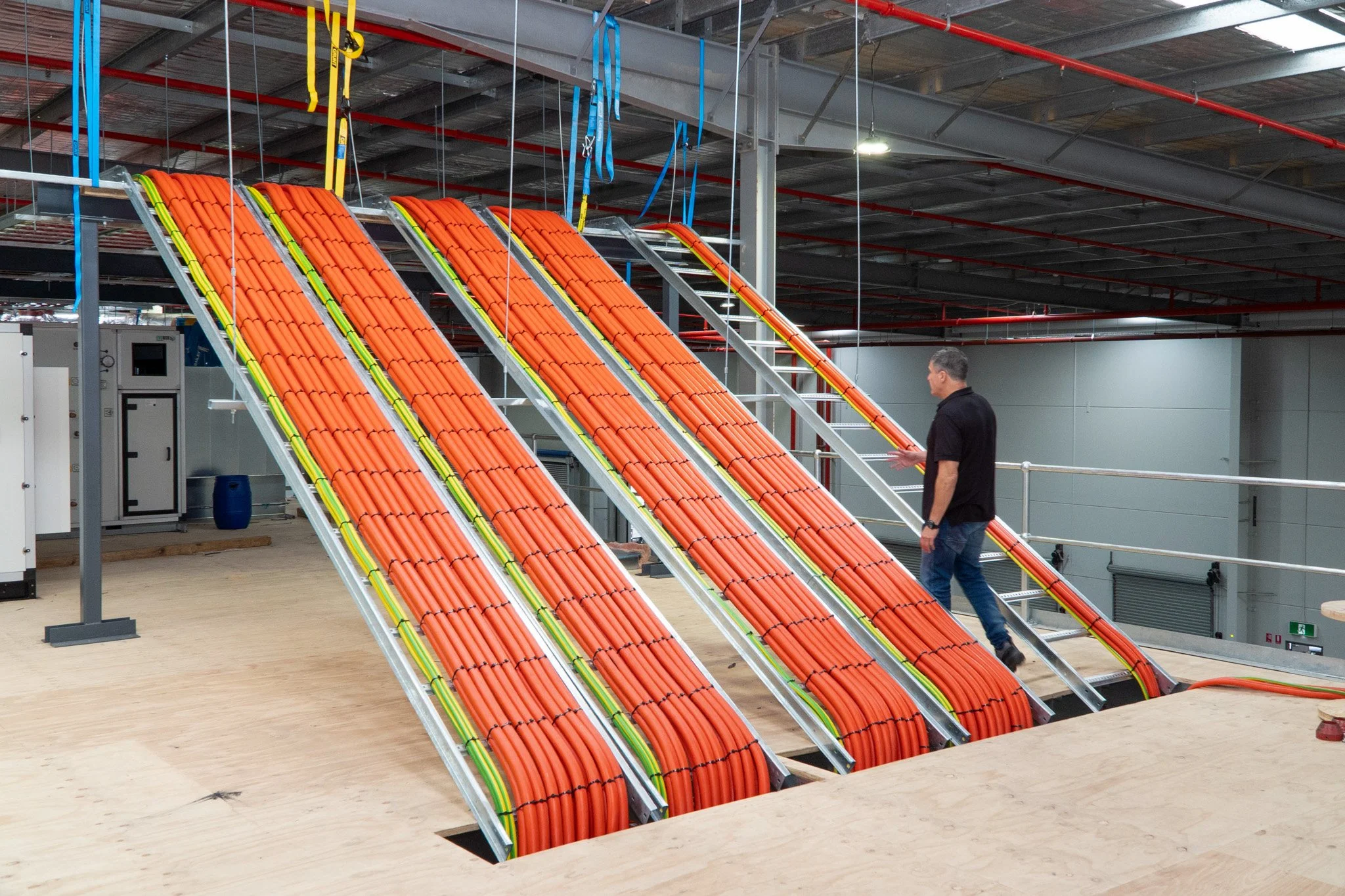 A person working on or inspecting orange cables on a large industrial setup inside a building with a high ceiling and metal framework.
