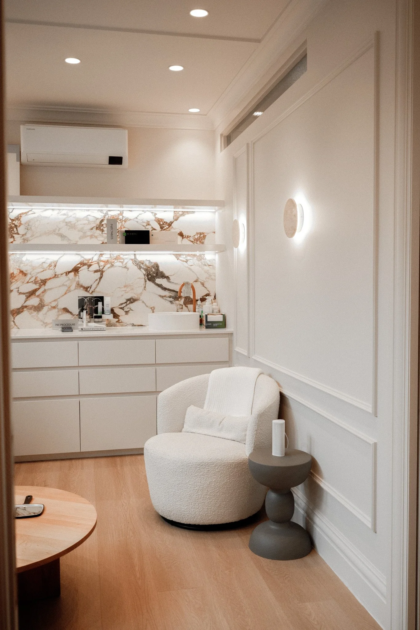 Interior of a modern room with light-colored walls and wooden flooring, featuring a curved white upholstered armchair, a small round side table with a white item on top, and a wooden coffee table. In the background, there is a white cabinet and a workspace with a marble-patterned backsplash, bottles, and skincare products. The room has recessed lighting, decorative wall sconces, a white air conditioning unit, and a long slim window near the ceiling.