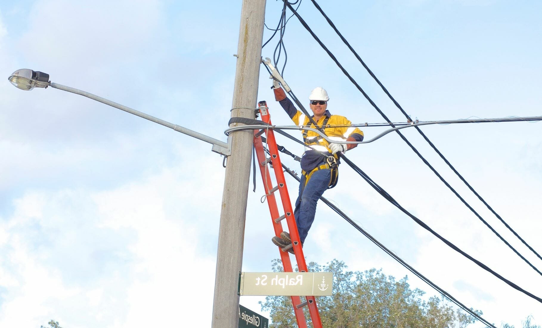 A specialised ASP Electrical worker standing on a ladder on a power pole, wearing safety gear, fixing or inspecting power lines with a cloudy sky background.