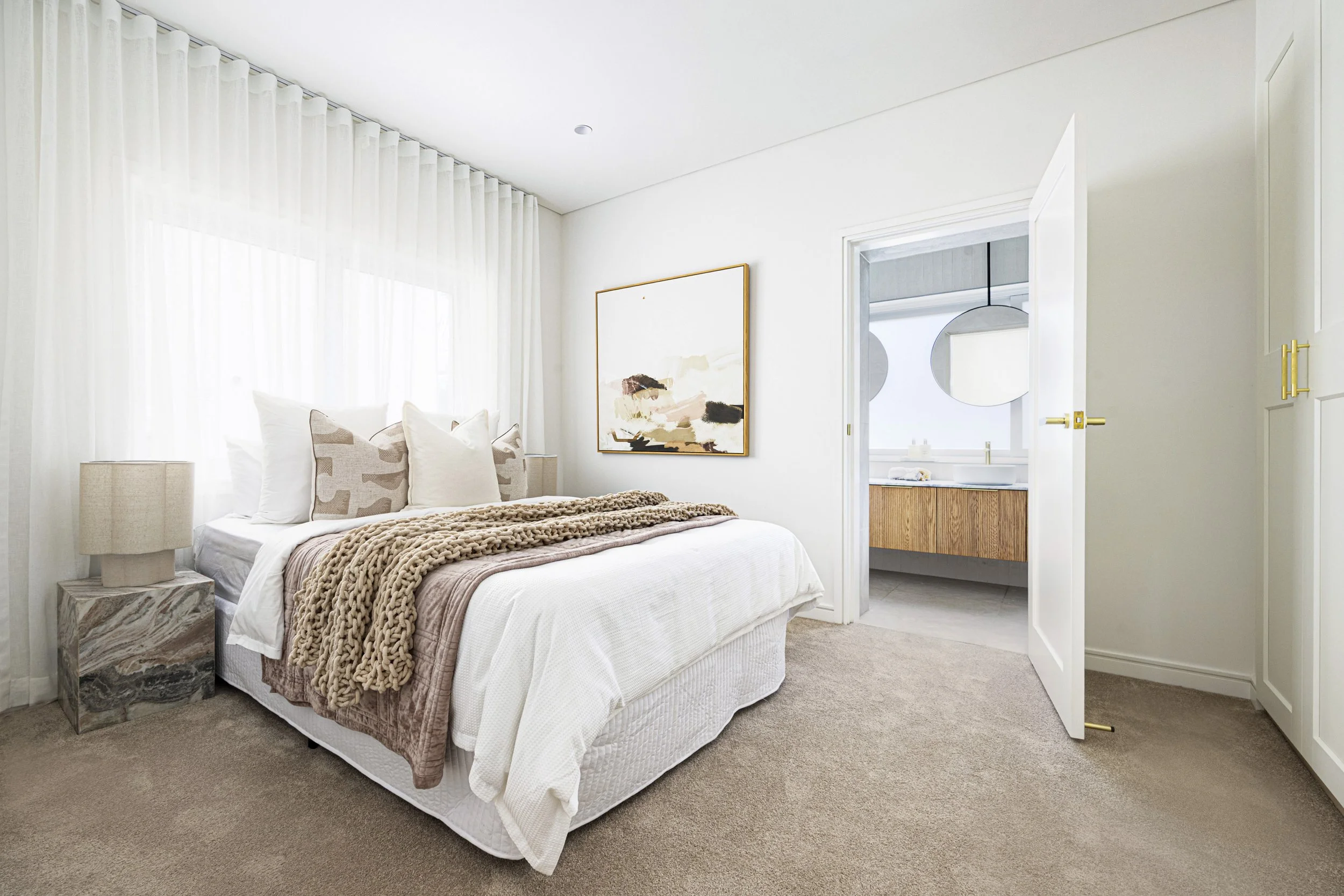 A bedroom with a bed, bedside tables, lamps, a wall art, and an open door leading to a bathroom with a sink and mirror.