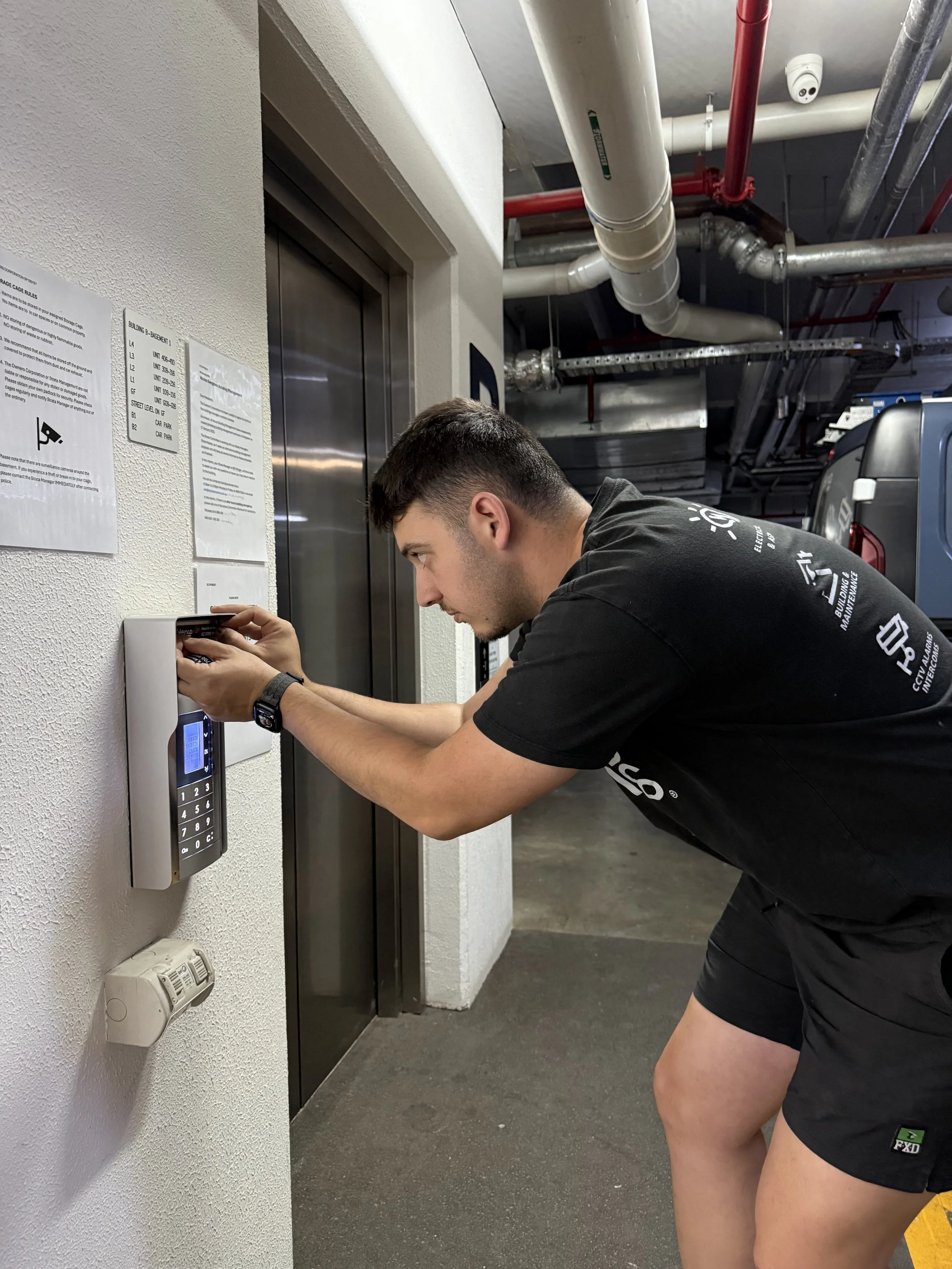 A man in black shorts and a black T-shirt is bent over near an elevator, entering a code or information into a keypad on the wall. The elevator door is closed, and there are information and safety notices posted on the wall next to it. Pipes and ventilation ducts are visible on the ceiling.