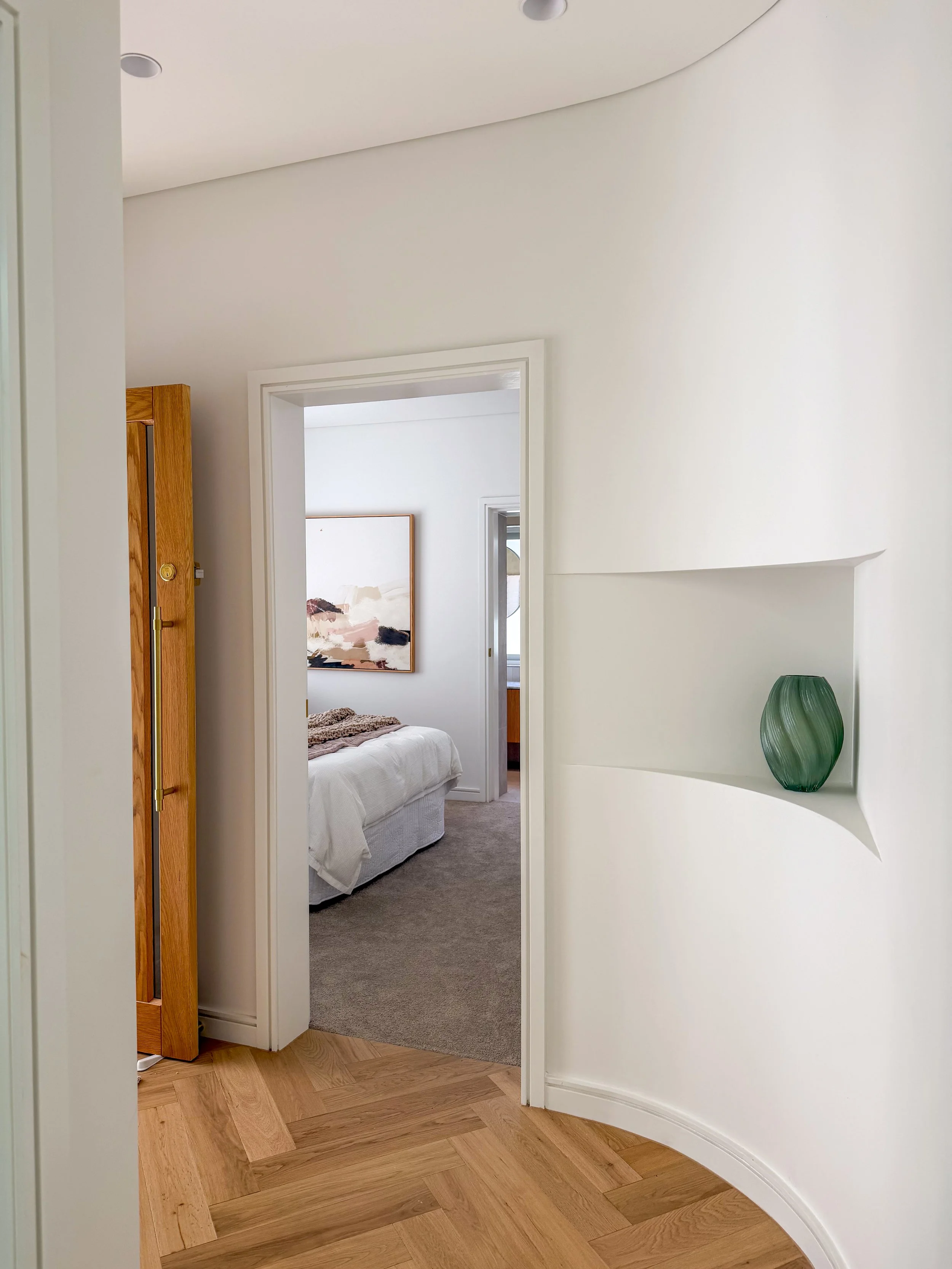 A view down a hallway into a bedroom with a bed, artwork on the wall, and a carpeted floor. The hallway has a curved wall with a green vase on a shelf and wooden flooring.