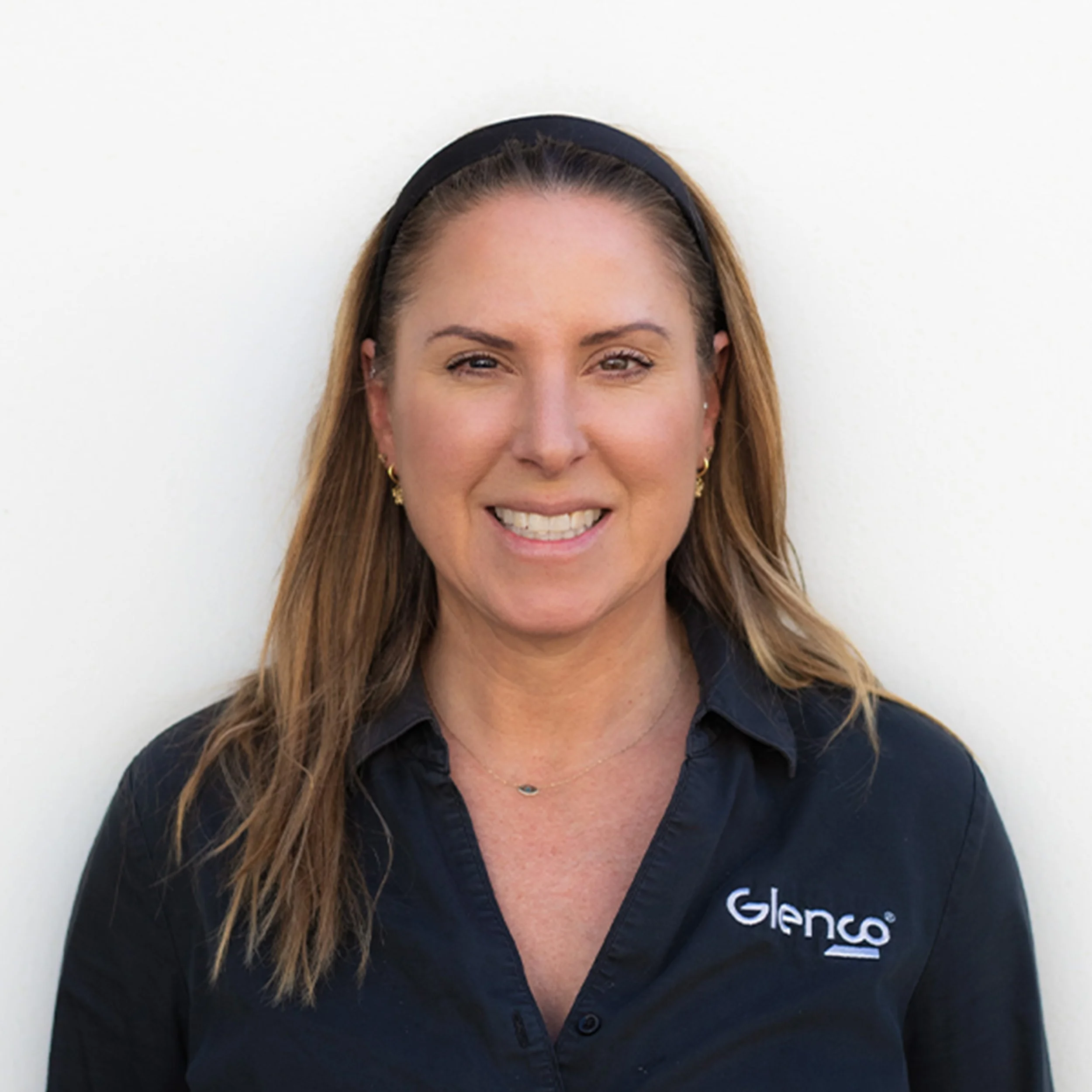 Close-up of a woman's face smiling, wearing a black headband and a black shirt with a logo, against a white background.