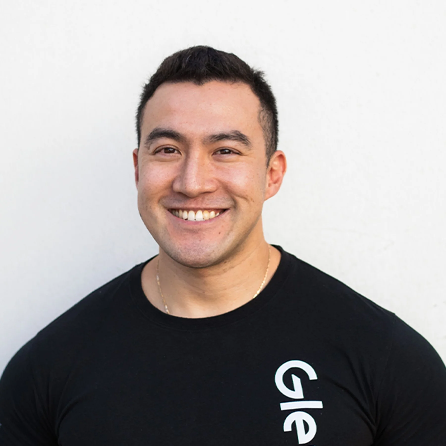 A smiling young man with short dark hair wearing a black T-shirt with partial white text on it, standing against a plain white background.