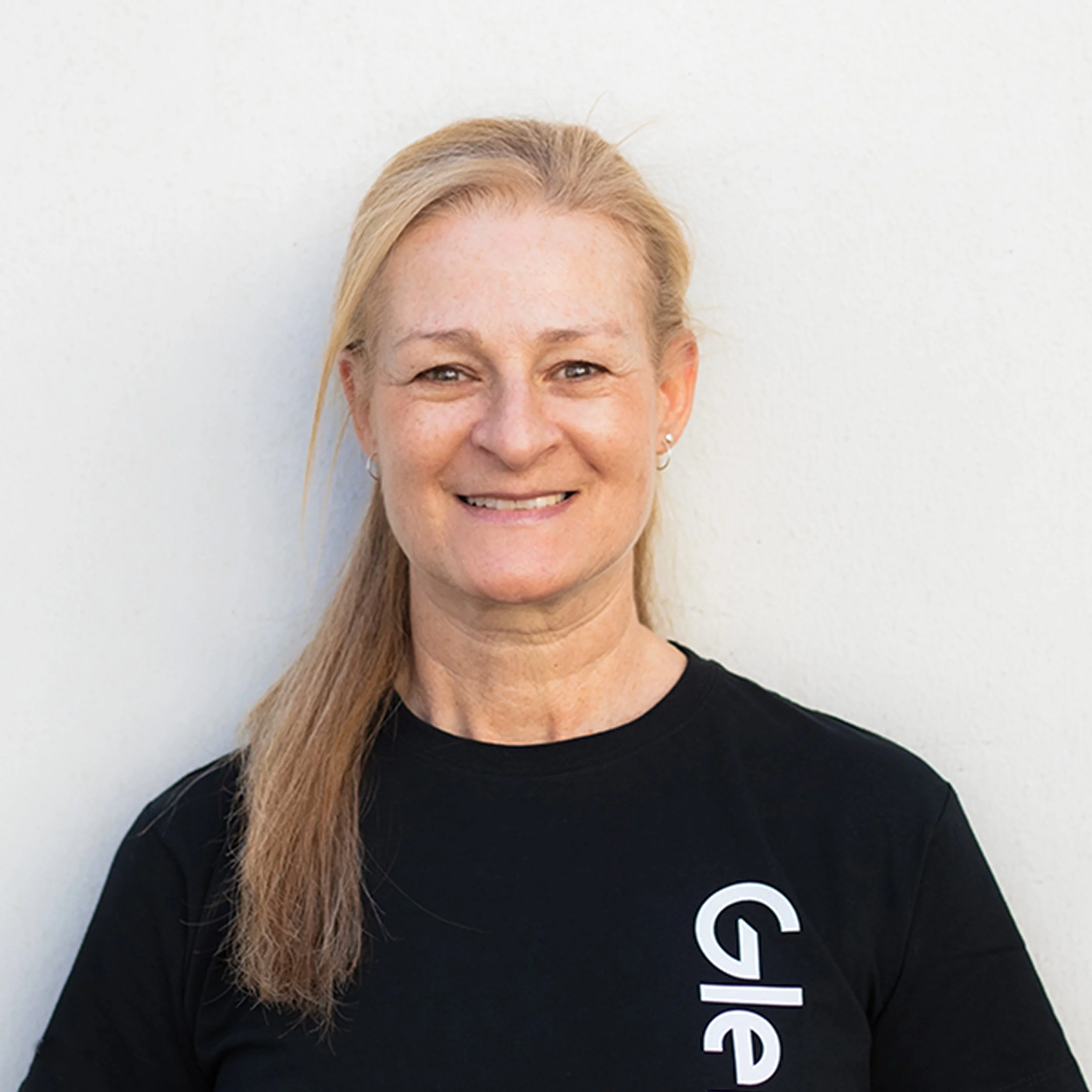 Close-up of a smiling woman with blonde hair, wearing earrings and a black T-shirt with white text, against a plain light background.