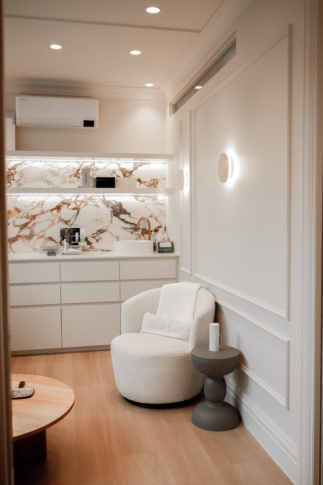 A cozy waiting area with a white textured armchair, a small round side table with a white object on top, and a wooden coffee table. The room features white walls with decorative molding, two wall-mounted lights, and a corner with a kitchen or vanity area that has a marble backsplash and various items on the counter. There is an air conditioner mounted near the ceiling and soft recessed lighting.