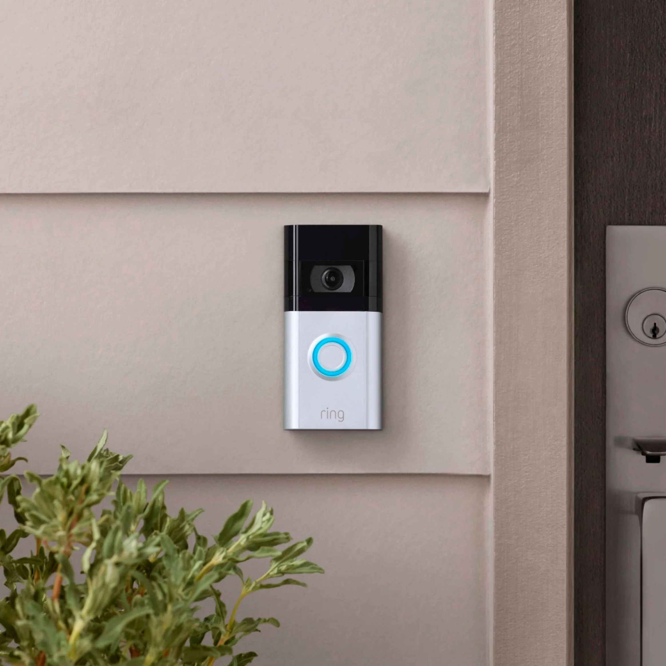 A Ring doorbell with a camera and a blue-lit button mounted on a white wall near a dark door.