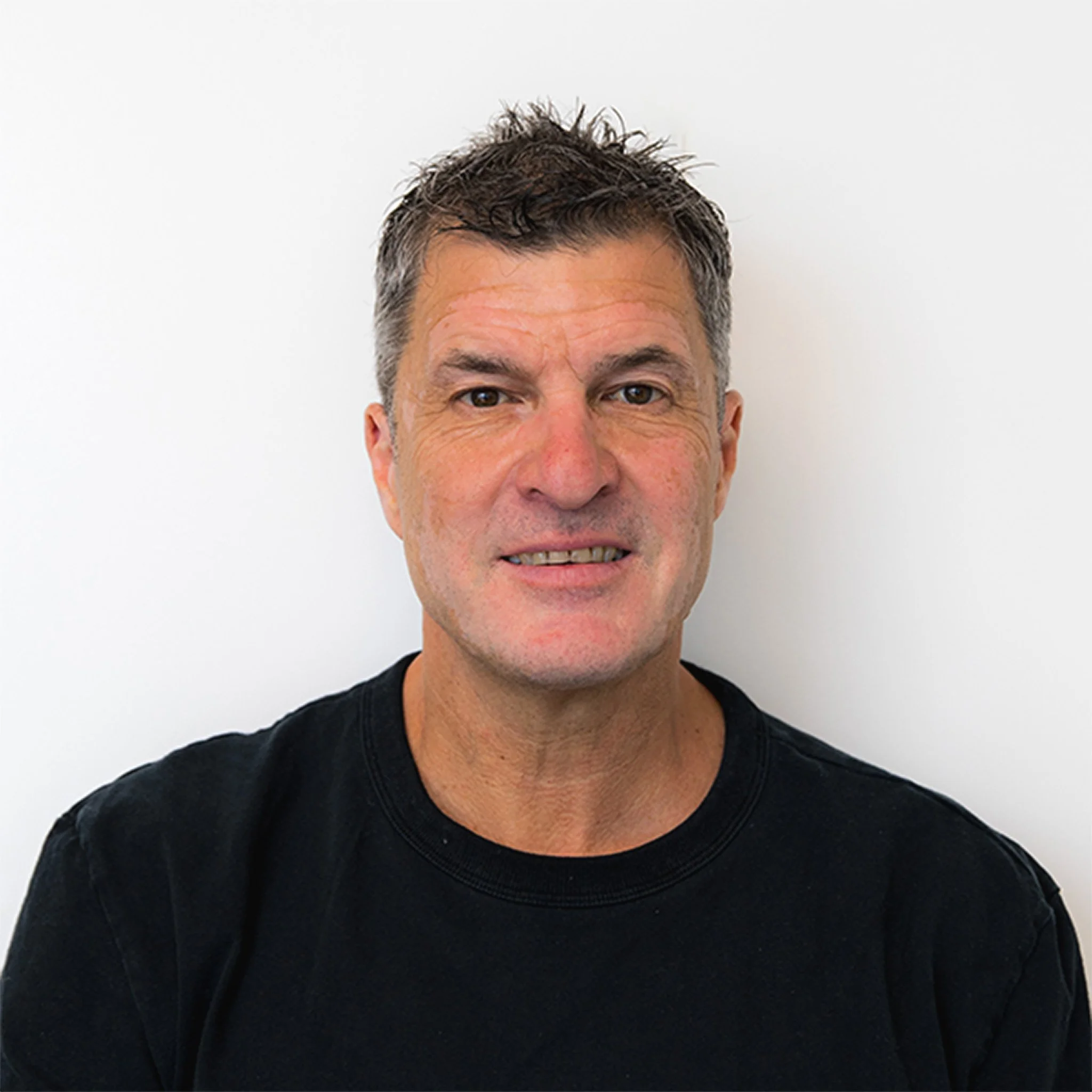 Close-up of man with short, slightly curly hair, smiling while wearing a black Glenco shirt against a plain white wall.