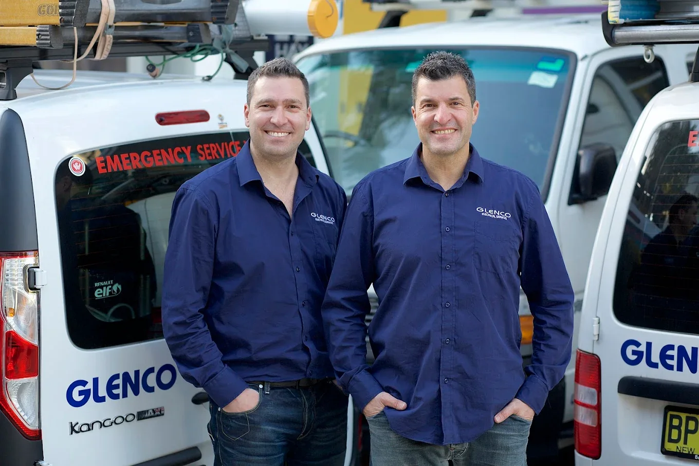 Two men in blue company shirts stand side by side in front of work vans with company logos, smiling at the camera.